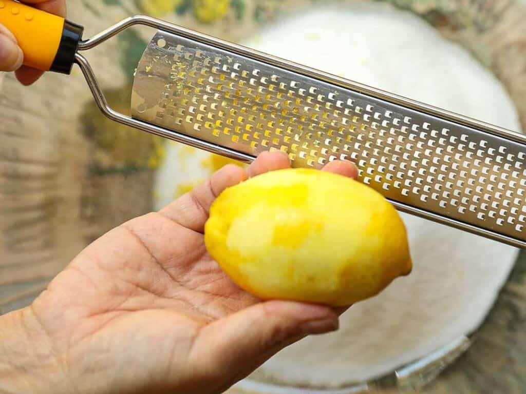 Close-up of hands holding a lemon and a zester with yellow handle. Lemon peel is partially grated. A white bowl is visible underneath, catching the lemon zest.